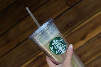 A hand holding a clear plastic tumbler with a green and white logo featuring a siren. The tumbler has a lid and straw, and it is held against a wooden, textured background.