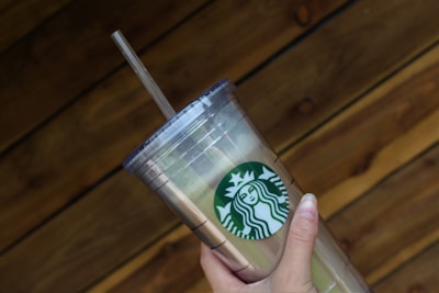 A hand holding a clear plastic tumbler with a green and white logo featuring a siren. The tumbler has a lid and straw, and it is held against a wooden, textured background.