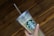 A hand holding a clear plastic tumbler with a green and white logo featuring a siren. The tumbler has a lid and straw, and it is held against a wooden, textured background.