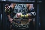 A muscular person wearing a sleeveless shirt with the words 'FULL POWER' is preparing for a weightlifting session. They have tattoos on their arms and are using lifting straps. The attire includes a leopard print weightlifting belt, and they are in a gym setting.