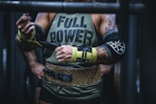 A muscular person wearing a sleeveless shirt with the words 'FULL POWER' is preparing for a weightlifting session. They have tattoos on their arms and are using lifting straps. The attire includes a leopard print weightlifting belt, and they are in a gym setting.