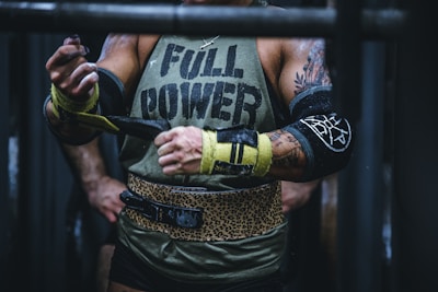 A muscular person wearing a sleeveless shirt with the words 'FULL POWER' is preparing for a weightlifting session. They have tattoos on their arms and are using lifting straps. The attire includes a leopard print weightlifting belt, and they are in a gym setting.