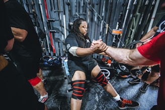 A person is seated and preparing for a workout, wearing knee wraps and surrounded by gym equipment. They are interacting with another person who is extending their hand in support. Several barbells and weightlifting gear are visible in the background.