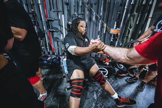A person is seated and preparing for a workout, wearing knee wraps and surrounded by gym equipment. They are interacting with another person who is extending their hand in support. Several barbells and weightlifting gear are visible in the background.