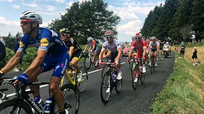 A group of cyclists are competing on a paved road surrounded by lush greenery and trees. They are wearing colorful jerseys and helmets, riding in a pack during what appears to be a professional road race. A few spectators are visible by the roadside, and a support vehicle follows the cyclists.