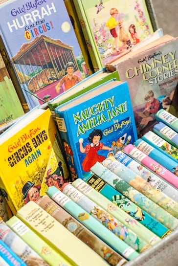 A collection of colorful children's books with illustrated covers, prominently featuring titles by Enid Blyton. The books are neatly arranged in a box, showcasing a variety of themes, including circus, adventure, and fantasy.