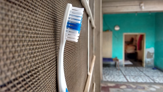 A toothbrush with blue and white bristles is positioned upright against a textured surface, possibly a mesh or a screen. In the background, an indoor setting with turquoise walls and a partially visible room with a tiled floor can be seen.
