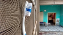 A toothbrush with blue and white bristles is positioned upright against a textured surface, possibly a mesh or a screen. In the background, an indoor setting with turquoise walls and a partially visible room with a tiled floor can be seen.