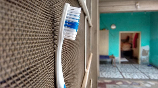 A toothbrush with blue and white bristles is positioned upright against a textured surface, possibly a mesh or a screen. In the background, an indoor setting with turquoise walls and a partially visible room with a tiled floor can be seen.