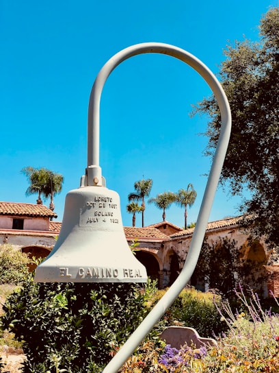 A large mission-style bell labeled 'El Camino Real' is mounted on a curved pole against a bright blue sky. In the background, there are red-tiled roof buildings with archways, typical of Spanish mission architecture. The area is surrounded by lush greenery, colorful flowers, and palm trees.