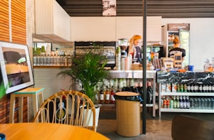 A cozy caf&eacute; interior with a counter displaying an assortment of bottles and snacks. Two people are working behind the counter, one of whom wears a shirt with 'SURFING.' A small plant is on the counter next to a coffee machine. A framed picture leans against the wall on a small side table, and there is some seating with wooden chairs and a table in the foreground.