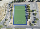 An aerial view of a rectangular sports field with a green synthetic surface, surrounded by a dark blue border. The field features marked lines indicating zones and goals. It is enclosed by a parking lot with multiple parking spaces, trees along the edges, and access roads with visible road markings. The area is open and surrounded by some greenery.