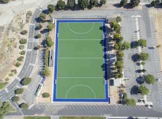 An aerial view of a rectangular sports field with a green synthetic surface, surrounded by a dark blue border. The field features marked lines indicating zones and goals. It is enclosed by a parking lot with multiple parking spaces, trees along the edges, and access roads with visible road markings. The area is open and surrounded by some greenery.