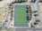 An aerial view of a rectangular sports field with a green synthetic surface, surrounded by a dark blue border. The field features marked lines indicating zones and goals. It is enclosed by a parking lot with multiple parking spaces, trees along the edges, and access roads with visible road markings. The area is open and surrounded by some greenery.
