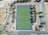 An aerial view of a rectangular sports field with a green synthetic surface, surrounded by a dark blue border. The field features marked lines indicating zones and goals. It is enclosed by a parking lot with multiple parking spaces, trees along the edges, and access roads with visible road markings. The area is open and surrounded by some greenery.