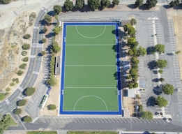 An aerial view of a rectangular sports field with a green synthetic surface, surrounded by a dark blue border. The field features marked lines indicating zones and goals. It is enclosed by a parking lot with multiple parking spaces, trees along the edges, and access roads with visible road markings. The area is open and surrounded by some greenery.