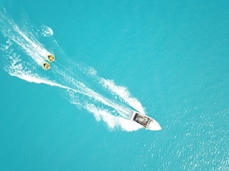 A speedboat races across a bright turquoise ocean, leaving white frothy trails in the water. Two yellow inflatable tubes are being towed behind the boat, with passengers enjoying the ride.