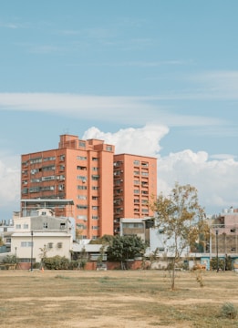 A large, red-brick residential building stands prominently against a backdrop of fluffy white clouds and clear blue sky. Surrounding the building are smaller, industrial-style structures with flat roofs, situated in a grassy, open area. Sparse trees and vegetation dot the landscape, contributing to a sense of urban tranquility.
