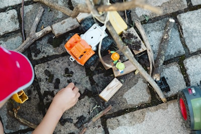 A child is playing with toy construction vehicles and wooden blocks on a cobblestone surface. There are sticks and dirt scattered around, and the child is engaging with the toys, possibly building or arranging something.