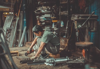 A man is sitting on the ground in a workshop, using a power tool to work on a metal piece. The area is cluttered with various tools, metal bars, and machinery. Shelves filled with paint cans and equipment are visible in the background.