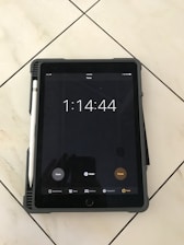 A tablet displaying a timer set for one hour, fourteen minutes, and forty-four seconds. The tablet has a pen attached to its side and is resting on a tiled floor. The screen shows options like 'Done,' 'Radar,' and 'Pause,' with icons for 'World Clock,' 'Alarm,' 'Bedtime,' 'Stopwatch,' and 'Timer' at the bottom.