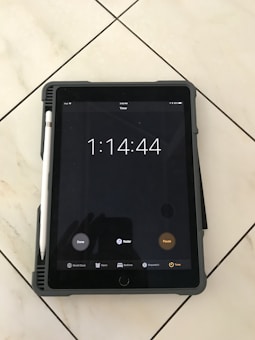 A tablet displaying a timer set for one hour, fourteen minutes, and forty-four seconds. The tablet has a pen attached to its side and is resting on a tiled floor. The screen shows options like 'Done,' 'Radar,' and 'Pause,' with icons for 'World Clock,' 'Alarm,' 'Bedtime,' 'Stopwatch,' and 'Timer' at the bottom.
