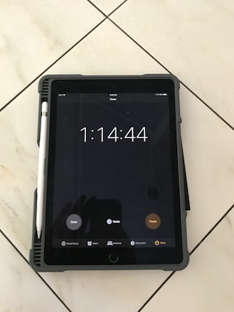 A tablet displaying a timer set for one hour, fourteen minutes, and forty-four seconds. The tablet has a pen attached to its side and is resting on a tiled floor. The screen shows options like 'Done,' 'Radar,' and 'Pause,' with icons for 'World Clock,' 'Alarm,' 'Bedtime,' 'Stopwatch,' and 'Timer' at the bottom.
