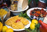 A vibrant assortment of Italian food items including fresh pasta, cheese, peppers, lemons, and a bottle of wine. The pasta is neatly arranged on a white plate, garnished with a single red tomato and parsley. Nearby is another plate filled with roasted red peppers and eggplants, alongside a variety of cheese and provisions. A wine bottle with a sealed cork and colorful label completes the arrangement, hinting at a rustic culinary setting.