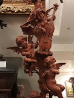 A detailed wooden sculpture of cherubic figures playing musical instruments. The figures are intricately carved with wings and are positioned in a playful arrangement. Various instruments like a cello, lute, and flute are being played by the cherubs. The sculpture is set against an ornately decorated background and appears to be part of a classical interior setting.