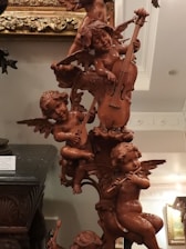 A detailed wooden sculpture of cherubic figures playing musical instruments. The figures are intricately carved with wings and are positioned in a playful arrangement. Various instruments like a cello, lute, and flute are being played by the cherubs. The sculpture is set against an ornately decorated background and appears to be part of a classical interior setting.