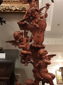 A detailed wooden sculpture of cherubic figures playing musical instruments. The figures are intricately carved with wings and are positioned in a playful arrangement. Various instruments like a cello, lute, and flute are being played by the cherubs. The sculpture is set against an ornately decorated background and appears to be part of a classical interior setting.