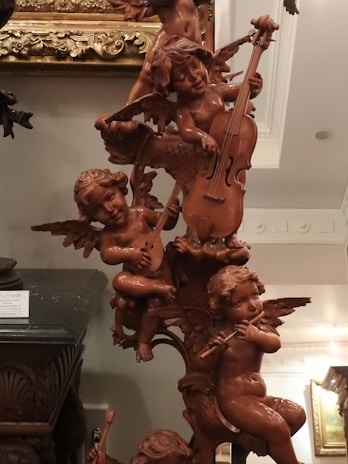 A detailed wooden sculpture of cherubic figures playing musical instruments. The figures are intricately carved with wings and are positioned in a playful arrangement. Various instruments like a cello, lute, and flute are being played by the cherubs. The sculpture is set against an ornately decorated background and appears to be part of a classical interior setting.