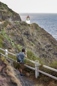 A person with a backpack is taking a photograph with a camera while standing on a trail along a rocky coastline. A white lighthouse with a red top is perched on the edge of a cliff overlooking the ocean.