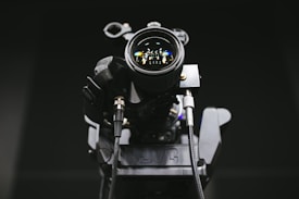A close-up view of a professional video camera with detailed focus on the lens, showing reflections of lights. The camera has a robust and technical appearance, with visible cables and mechanical parts.