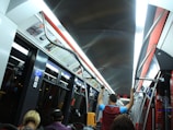 The interior of a modern subway or tram with overhead lighting, metal handrails, and passengers seated and standing. Advertisements are visible above the windows, and there are several signs indicating various instructions. The environment appears clean and organized.