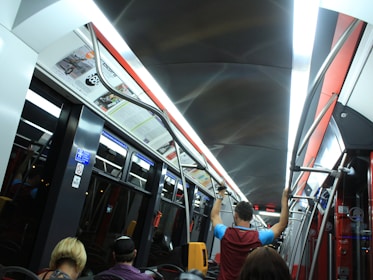 The interior of a modern subway or tram with overhead lighting, metal handrails, and passengers seated and standing. Advertisements are visible above the windows, and there are several signs indicating various instructions. The environment appears clean and organized.
