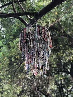 A collection of rosary beads, featuring numerous crosses and beads in various colors, hangs from the branch of a tree. The background is filled with lush green leaves, providing a serene natural setting.
