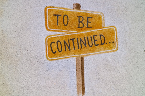 A wall features a painted design of a wooden post with two rectangular, orange signs resembling street signs. These signs display the text 'TO BE' on the top sign and 'CONTINUED...' on the bottom sign, with black lettering.