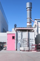 An industrial facility with a tall exhaust stack protruding from the rooftop of a rectangular structure. The structure has a pink door on the left and a smaller pink section on the right. There are metal ladders attached to the side and various industrial fixtures around the building. Adjacent structures and the clear blue sky are visible in the background.