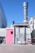 An industrial facility with a tall exhaust stack protruding from the rooftop of a rectangular structure. The structure has a pink door on the left and a smaller pink section on the right. There are metal ladders attached to the side and various industrial fixtures around the building. Adjacent structures and the clear blue sky are visible in the background.
