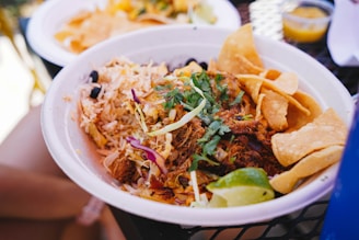 A bowl filled with a variety of Mexican-inspired ingredients, including shredded meat, rice, sliced avocado, tortilla chips, and garnished with fresh cilantro and lime wedges. The dish is presented on a table with another plate and a small container of sauce in the background.