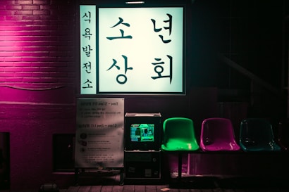 A brightly lit Korean sign is displayed against a brick wall with neon lighting. Below the sign are several colorful plastic chairs in green, purple, and blue. A small machine, possibly a vending or arcade machine, is positioned underneath the sign.