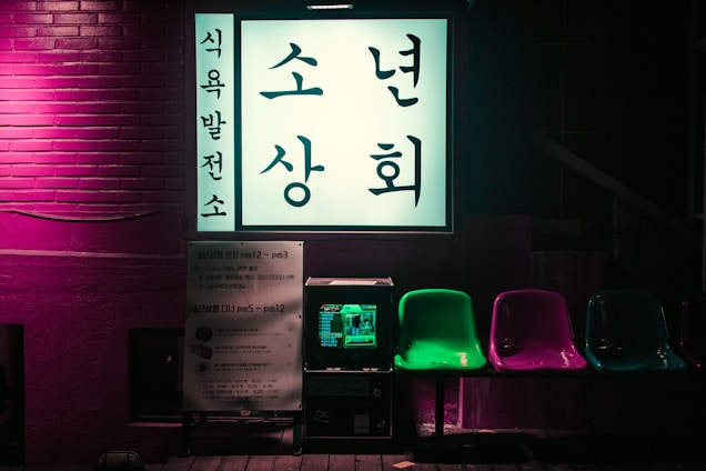 A brightly lit Korean sign is displayed against a brick wall with neon lighting. Below the sign are several colorful plastic chairs in green, purple, and blue. A small machine, possibly a vending or arcade machine, is positioned underneath the sign.