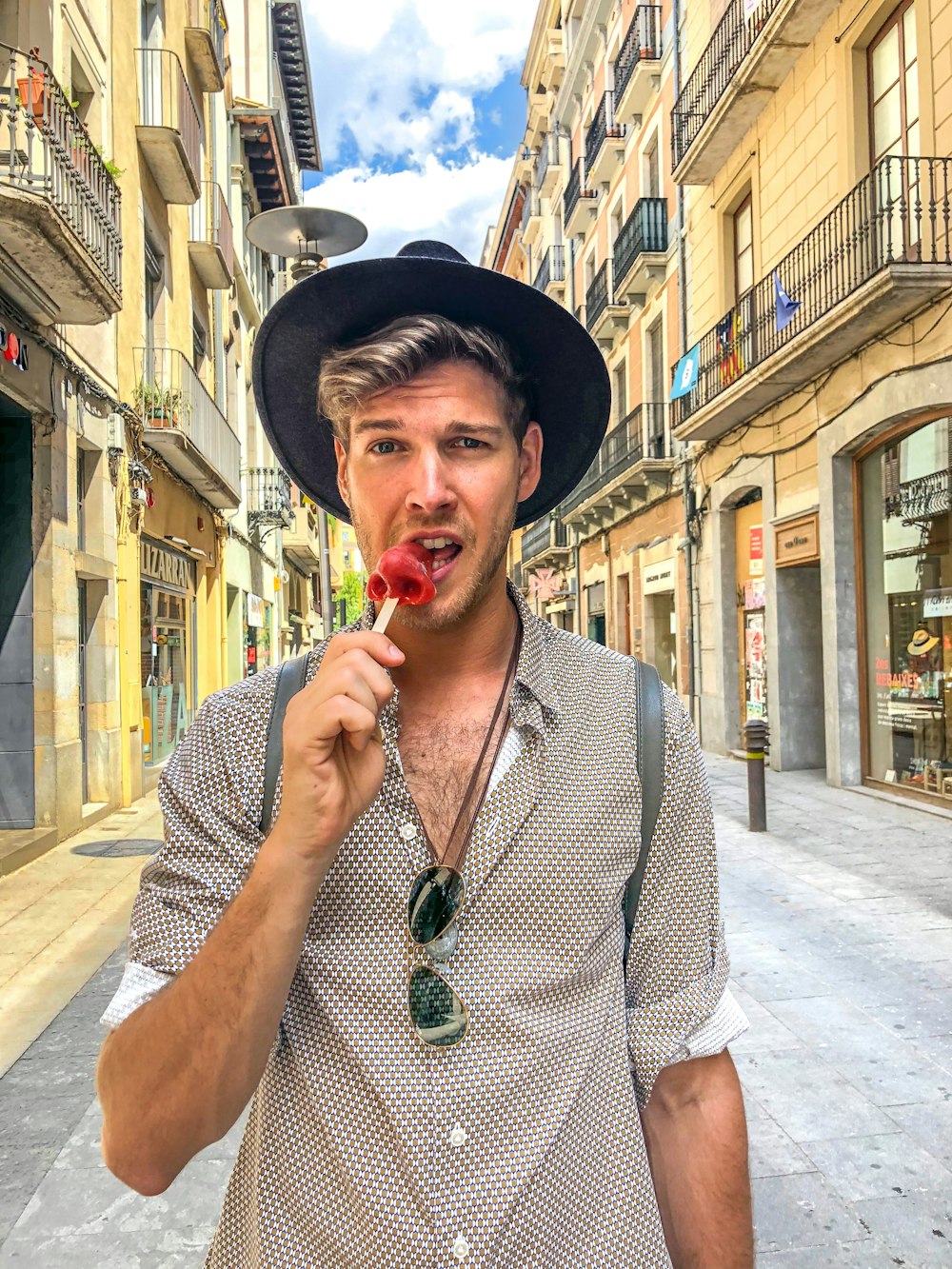 hombre con sombrero y camisa gris mordiendo caramelo rojo en un palo