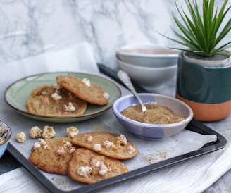 A tray with freshly baked cookies containing popcorn, placed on a piece of parchment paper. Beside the tray is a small bowl filled with brown sugar or spice, accompanied by a spoon. There's also a potted plant in a stylish, multicolored container, and a stack of ceramic bowls in the background.