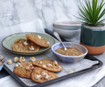A tray with freshly baked cookies containing popcorn, placed on a piece of parchment paper. Beside the tray is a small bowl filled with brown sugar or spice, accompanied by a spoon. There's also a potted plant in a stylish, multicolored container, and a stack of ceramic bowls in the background.