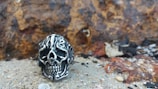 A metallic skull ring with intricate engravings sits on rocky terrain. The background features a textured, rust-colored rock surface.