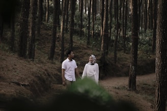 A man and a woman walk together in a tranquil forest setting, surrounded by tall trees with straight trunks. The forest floor is a path covered in brown earth and pine needles. The woman wears a white outfit and a hijab, while the man is dressed in a white short-sleeved shirt and khaki pants.