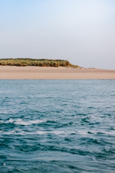 A serene seascape with gentle waves in the foreground and a sandy shore in the background, topped with green grass. The sky is clear with a soft gradient from light blue to white.