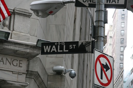 A street sign for Wall Street mounted on a pole, alongside a one-way traffic sign and a 'no right turn' sign. The background shows stone architecture and a partial view of the American flag, indicating a financial district setting.